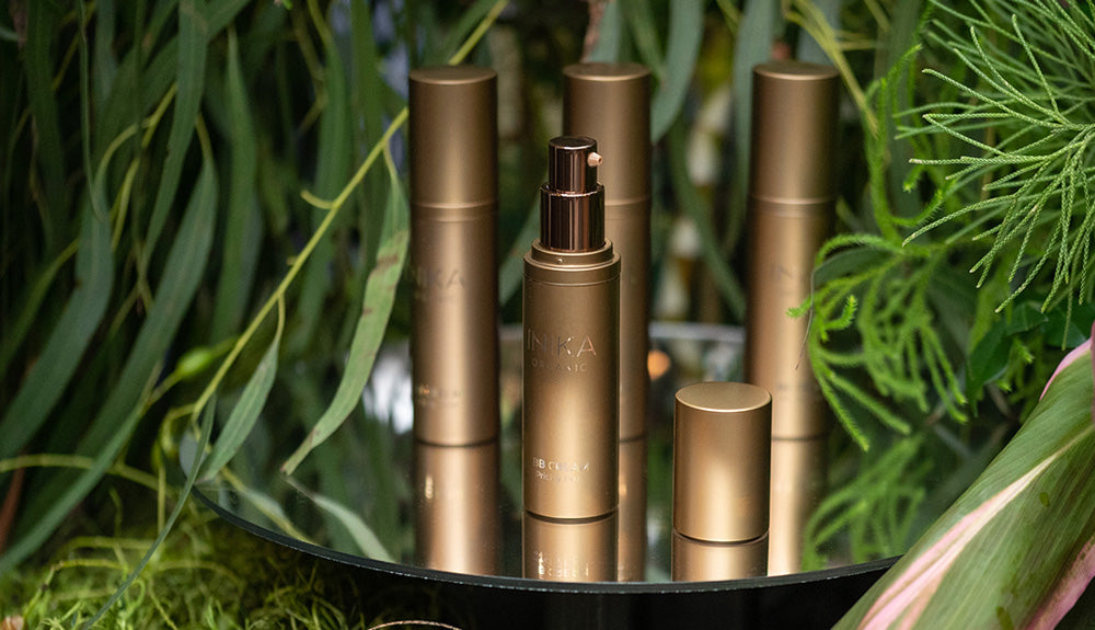 Luxurious gold skincare bottles displayed on a mirrored surface, surrounded by lush green foliage. Reflective and elegant composition.
